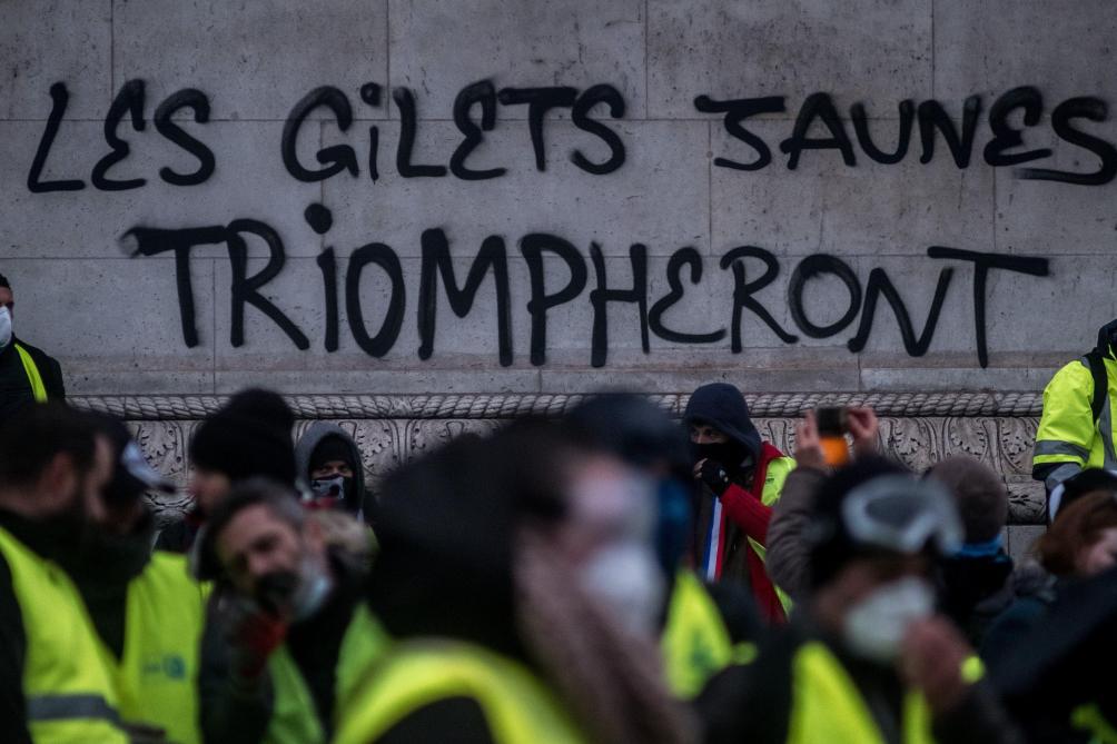 Sur un mur, le graffiti « Les gilets jaunes triompheront » derrière une foule en gilets jaunes